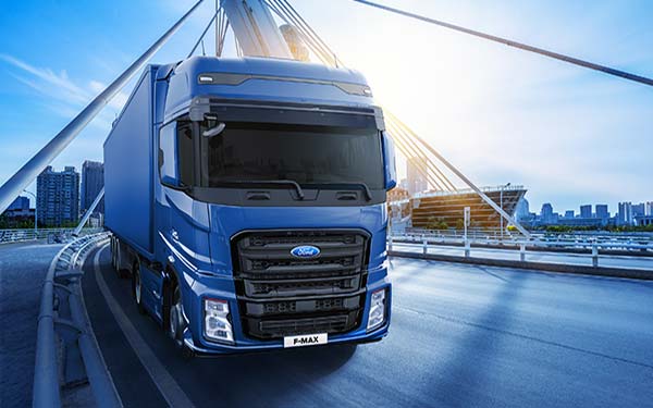 OsnaTruck_Verkauf_Ford-F-Max2_600x375pxl Blauer Ford Truck mit blauem Auflieger fährt über eine Stahl-Hängebrücke. Im Hintergrund sind Hochhäuser.