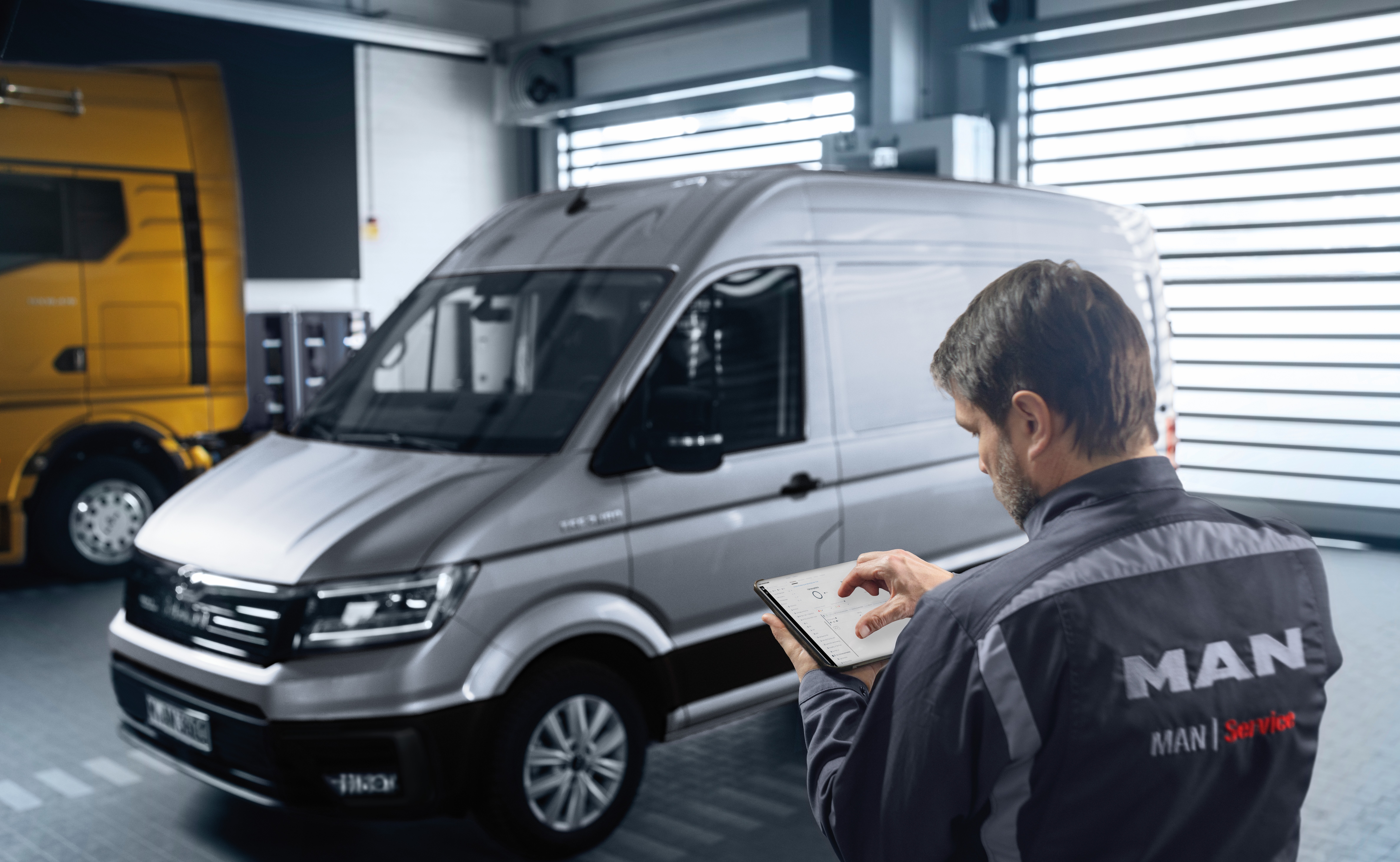 MAN TGE Next Level_Service_2 MAN Servicemitarbeiter steht mit dem Rücken ins Bild und Tablet in der Hand vor einem grauen TGE in der Werkstatthalle