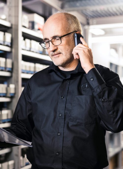 Teilelager Mann mit schwarzem Hemd, telefoniert, Teileregal im Hintergrund